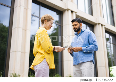 A woman in a yellow shirt hands money to a man in a blue shirt, both outdoors in front of a building. A woman in a yellow shirt hands money to a man in a blue shirt, both outdoors in front of a building. 129730222