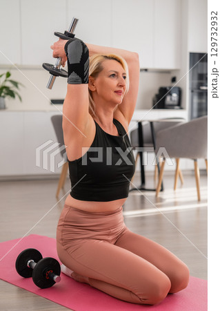 Focused blonde middle aged woman sitting on floor mat lifting dumbbell behind head to train triceps during home workout in modern living room Focused blonde middle aged woman sitting on floor mat lifting dumbbell behind head to train triceps during home workout in modern living room 129732932