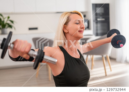 Focused blonde middle aged woman lifting dumbbells overhead to strengthen shoulder muscles during home workout in modern living room Focused blonde middle aged woman lifting dumbbells overhead to strengthen shoulder muscles during home workout in modern living room 129732944