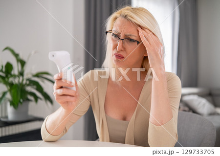 Blonde middle aged woman in beige outfit looking worried at thermometer, experiencing high temperature and discomfort in modern living room Blonde middle aged woman in beige outfit looking worried at thermometer, experiencing high temperature and discomfort in modern living room 129733705