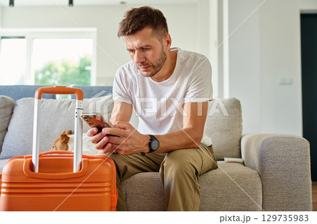 Man sitting on sofa with suitcase and using smartphone 129735983