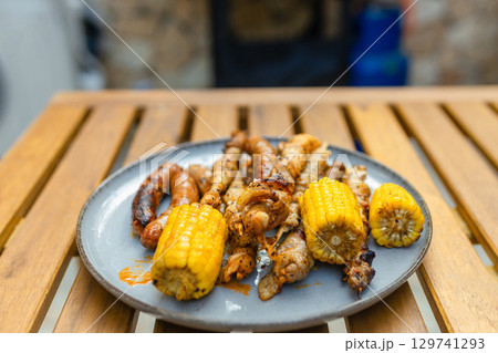 Grilled sausages, skewers, and corn on a plate near a gas barbecue with vegetables and flatbreads cooking. Outdoor summer cooking scene with delicious grilled food. Grilled sausages, skewers, and corn on a plate near a gas barbecue with vegetables and flatbreads cooking. Outdoor summer cooking scene with delicious grilled food. 129741293