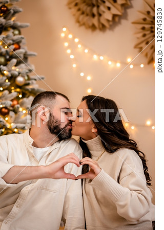 Couple Sharing a Kiss by Christmas Tree with Heart Gesture 129745830