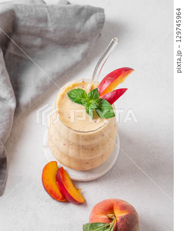 Smoothie with peach and banana in a glass on a light background with tropical fruits and napkin. Smoothie with peach and banana in a glass on a light background with tropical fruits and napkin. 129745964