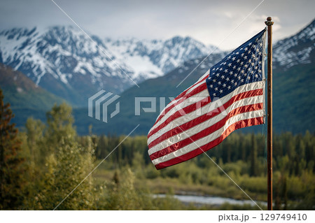 The US flag on a pole flutters in the wind against the backdrop of a clear blue sky and untouched nature with forest and mountains. 129749410