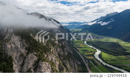 Aerial view of a valley in the Austrian Alps with low clouds covering the mountainside, featuring the Inn river, fields, and a small town 129749458
