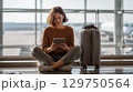 Young traveler reading on tablet while waiting at airport with suitcase nearby 129750564