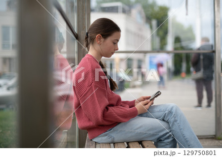 Young girl sitting at bus stop, scrolling through social media feed. Young girl sitting at bus stop, scrolling through social media feed. 129750801