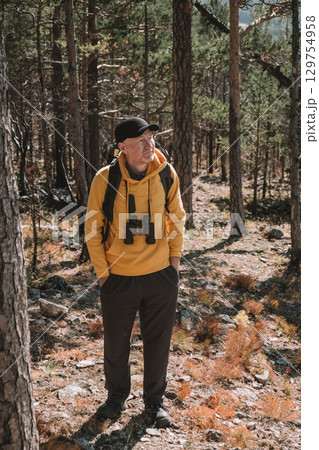 elderly man explores forest wearing yellow hoodie, examining nature binoculars during autumn. 129754958