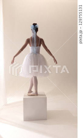 Elegant ballerina in white tutu with blue bow posing on pointe box stage 129755131