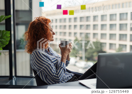 Side view of businesswoman resting in an office, holding a cup of coffee. 129755449
