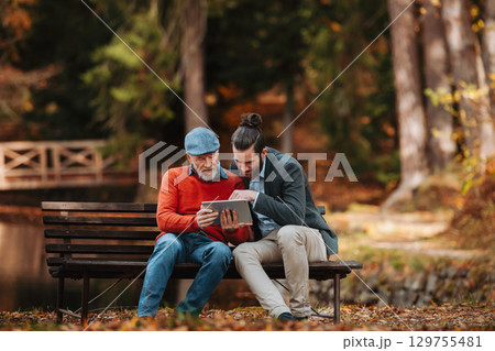 Senior father and his son sitting on bench by lake in nature, looking at tablet. 129755481