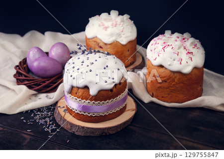 Easter cakes on a dark wooden background, decorated with lavender and marshmallows, and painted Easter eggs in a wicker nest. 129755847