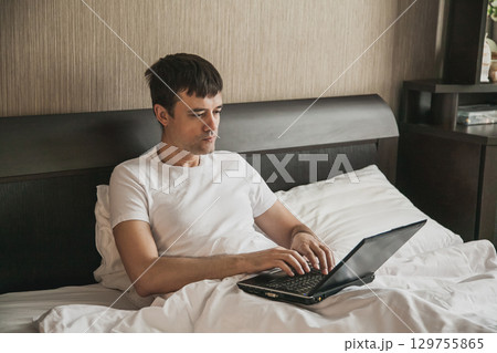 A middle-aged man is sitting in bed in his bedroom and working on a laptop A middle-aged man is sitting in bed in his bedroom and working on a laptop 129755865