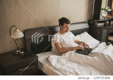A middle-aged man is sitting in bed in his bedroom and working on a laptop 129755866