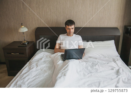 A middle-aged man is sitting in bed in his bedroom and working on a laptop A middle-aged man is sitting in bed in his bedroom and working on a laptop 129755873