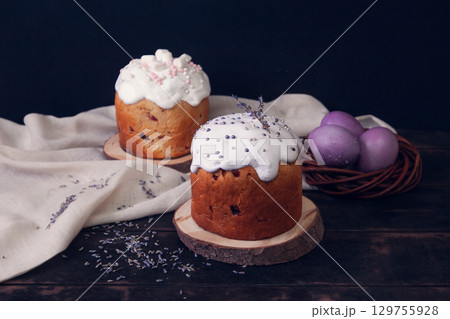 Easter cakes on a dark background, decorated with lavender and marshmallows, and painted Easter eggs in a wicker nest. 129755928