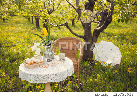 Romantic picnic in the spring garden. Decorations for a photo shoot in a blooming Apple orchard. A coffee table filled with cups, candy, pastries, and a vase of tulips, wicker chair, lace umbrella. 129756013
