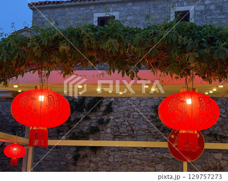 Red Chinese lanterns glowing in the evening street under green vines and stone wall. Tradition, culture and festive atmosphere symbolized by light and heritage decoration. 129757273