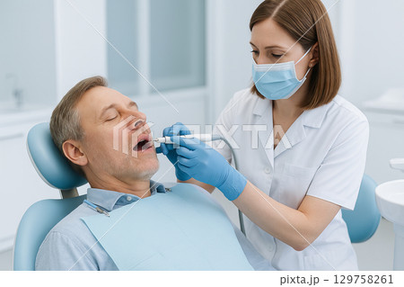 Female dentist performing dental procedure on mature male patient in modern clinic setting. concept of professional healthcare, dental care, oral hygiene 129758261