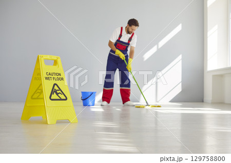 Caution wet floor sign with male young cleaner man mopping empty room or office in background. Caution wet floor sign with male young cleaner man mopping empty room or office in background. 129758800