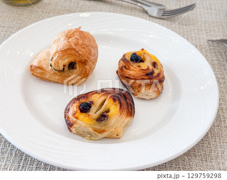 A white plate with croissant and raisin pastries served on a breakfast table. Sweetness, indulgence, hospitality and morning choice in hotel dining atmosphere. 129759298