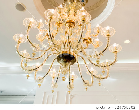 A golden chandelier with crystal ornaments hanging from a decorated ceiling. Elegance, luxury, atmosphere and architectural style in hotel interior and dining hall design. 129759302
