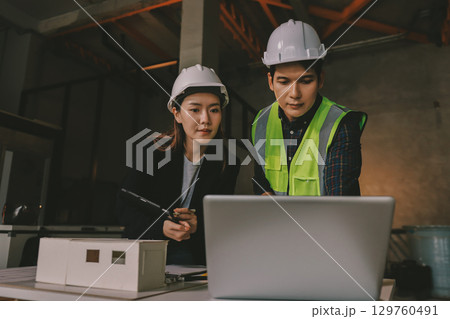 Construction manager and engineer dressed in orange work vests and hard helmets explore construction documentation on the building site near the steel frames Construction manager and engineer dressed in orange work vests and hard helmets explore construction documentation on the building site near the steel frames 129760491