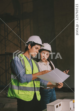 Construction manager and engineer dressed in orange work vests and hard helmets explore construction documentation on the building site near the steel frames 129760515