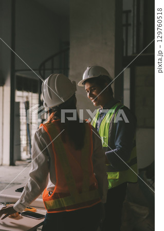 Construction manager and engineer dressed in orange work vests and hard helmets explore construction documentation on the building site near the steel frames Construction manager and engineer dressed in orange work vests and hard helmets explore construction documentation on the building site near the steel frames 129760518