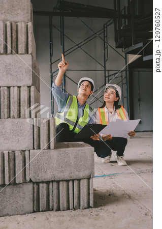 Construction manager and engineer dressed in orange work vests and hard helmets explore construction documentation on the building site near the steel frames 129760527