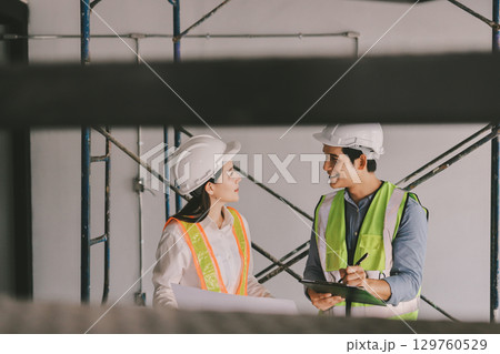 Construction manager and engineer dressed in orange work vests and hard helmets explore construction documentation on the building site near the steel frames Construction manager and engineer dressed in orange work vests and hard helmets explore construction documentation on the building site near the steel frames 129760529