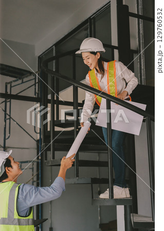 Construction manager and engineer dressed in orange work vests and hard helmets explore construction documentation on the building site near the steel frames Construction manager and engineer dressed in orange work vests and hard helmets explore construction documentation on the building site near the steel frames 129760532