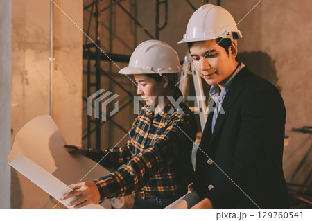 Construction manager and engineer dressed in orange work vests and hard helmets explore construction documentation on the building site near the steel frames 129760541