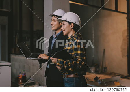 Construction manager and engineer dressed in orange work vests and hard helmets explore construction documentation on the building site near the steel frames 129760555