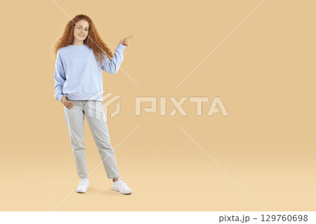 Young woman pointing to copy space and looking at camera with smiling face on a beige background. 129760698
