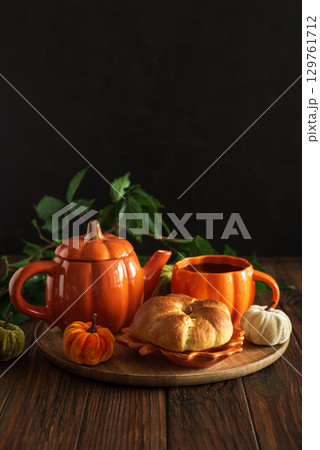 Autumnal tea set with pumpkin-shaped teapot and mug, and a baked bun on a wooden table. Autumnal tea set with pumpkin-shaped teapot and mug, and a baked bun on a wooden table. 129761712