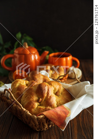 Autumnal tea set with baked buns into basket, pumpkin-shaped teapot and mug on a wooden table. 129761714