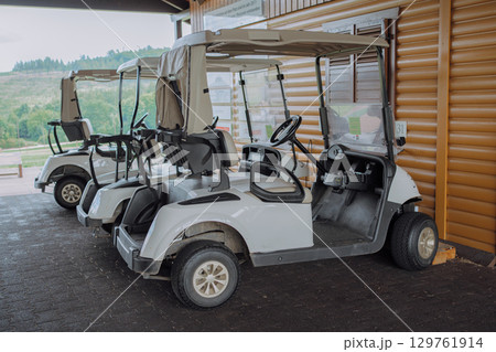 Golf carts are parked in a neat row under a clubhouse canopy. The vehicles are ready for golfers to use. The scene is serene with a glimpse of the golf course in the background. Golf carts are parked in a neat row under a clubhouse canopy. The vehicles are ready for golfers to use. The scene is serene with a glimpse of the golf course in the background. 129761914