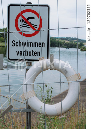 No swimming sign with a safety ring by a river, emphasizing caution. The sign prohibits swimming to ensure safety near the water. Vegetation surrounds the sign 129762536