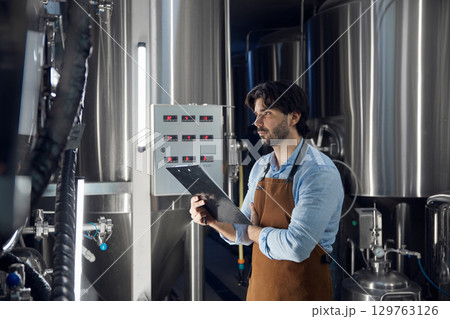 A man brewer in an apron stands confidently in a factory, arms crossed A man brewer in an apron stands confidently in a factory, arms crossed 129763126