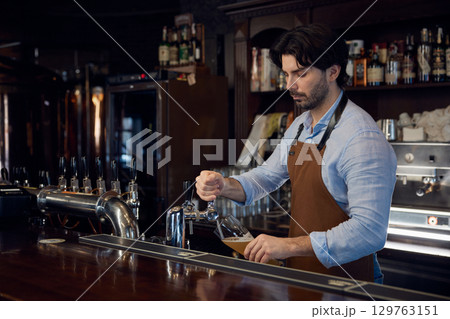A bartender skillfully pours a beer from a tap into a glass container A bartender skillfully pours a beer from a tap into a glass container 129763151