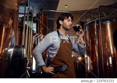 Brewer wearing an apron and is holding a glass filled with fresh beer Brewer wearing an apron and is holding a glass filled with fresh beer 129763153