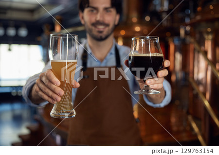 A man wearing an apron is expertly holding two glasses of beer 129763154