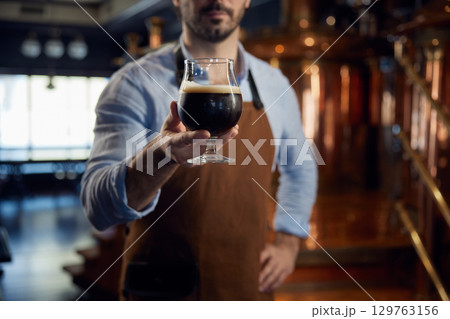 A man brewer wearing an apron and is holding a glass filled with beer 129763156