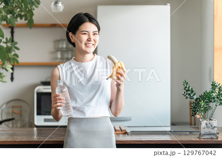 キッチンでバナナを食べるスポーツウェア姿の若いアジア人女性（トレーニング,エネルギー補給,間食） 129767042