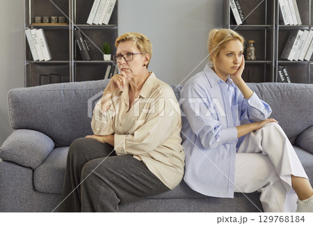 Mother and daughter home conflict, sitting back-to-back on sofa, senior adult and young woman upset Mother and daughter home conflict, sitting back-to-back on sofa, senior adult and young woman upset 129768184
