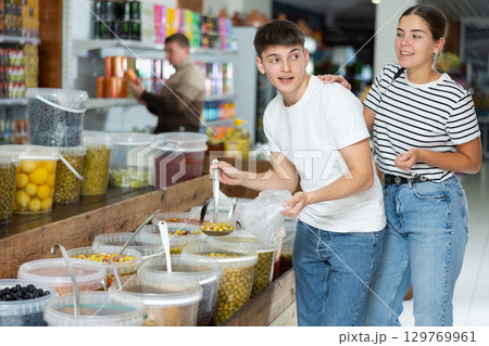 Young couple chooses marinated olives in supermarket, scooping them from bucket with ladle into plastic bag 129769961