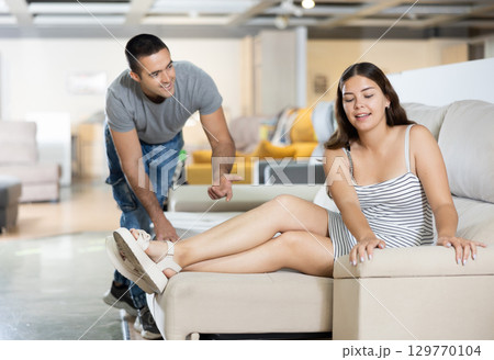 Couple chooses furniture in store.Wife is sitting on sofa,husband is standing next to her Couple chooses furniture in store.Wife is sitting on sofa,husband is standing next to her 129770104