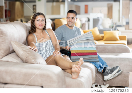 In store wife sit with legs on gray sofa to test in use,husband examines upholstery fabric 129770286